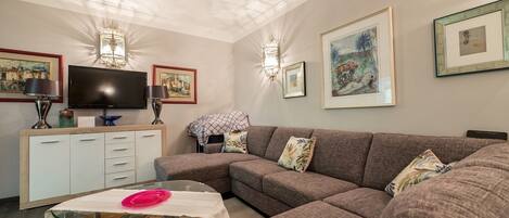 A living room with a sectional sofa, a glass coffee table, a flat-screen TV, a cabinet with drawers, and wall-mounted lights.