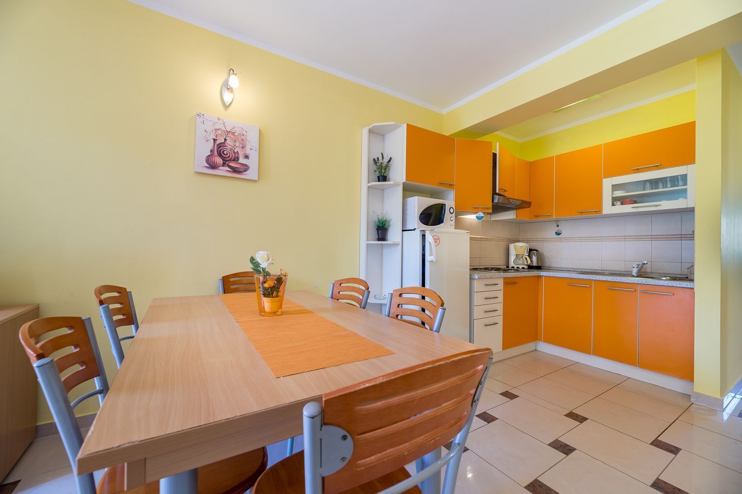 A compact kitchen with orange cabinets, a wooden dining table with chairs, and a small refrigerator.