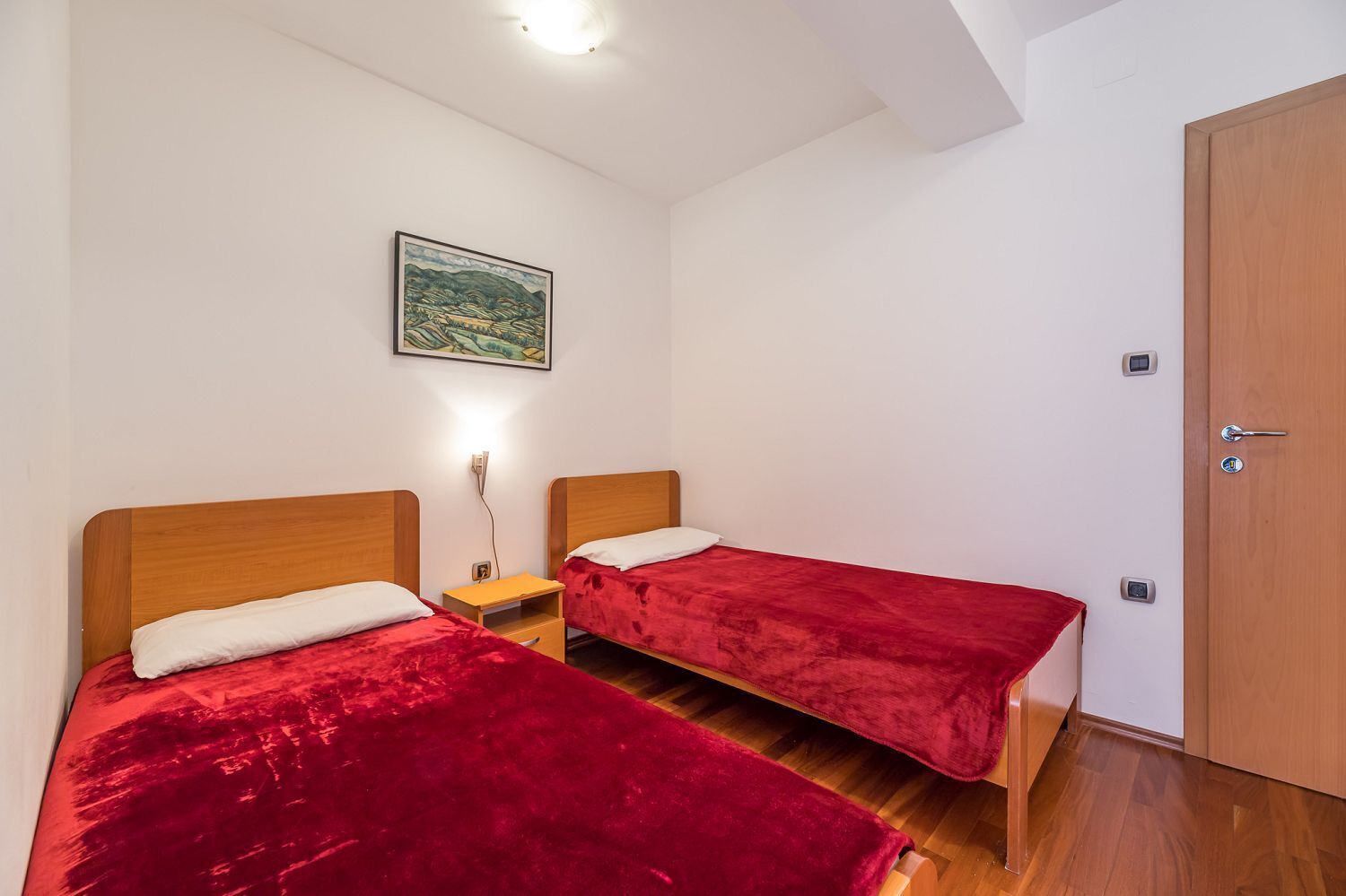 A room with two single beds, wooden headboards, a nightstand, a red blanket, a painting on the wall, and a wooden door.