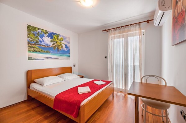 Un dormitorio con cama de madera, una manta roja, un cuadro de una escena de playa, una mesa de comedor y una silla.