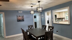 Un coin repas avec une table et des chaises en bois, un ventilateur de plafond et une vue sur la cuisine grâce à une porte ouverte.