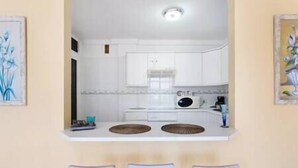 Una cocina compacta con armarios blancos, una estufa y un lavabo. Hay dos sillas frente a la encimera.