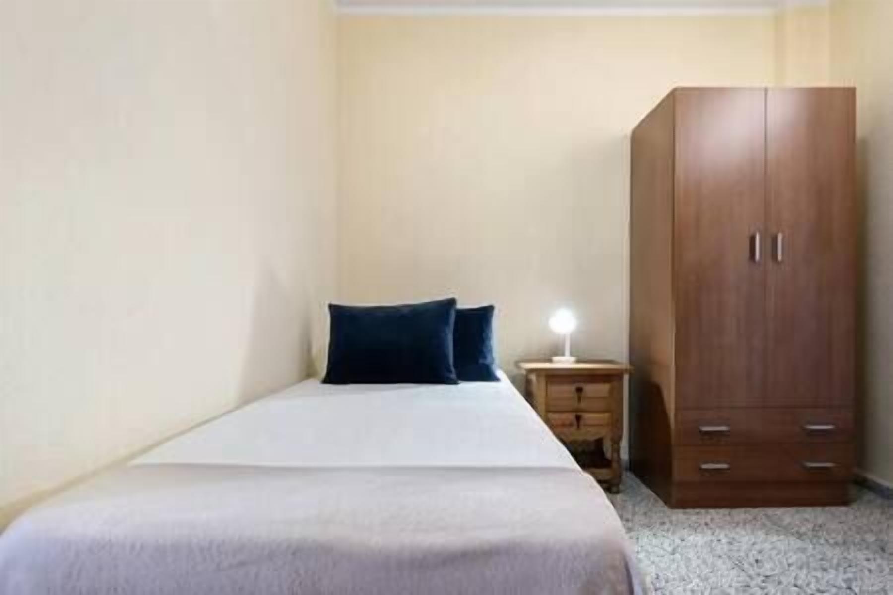 A small, simple room with a bed, a nightstand, and a wooden wardrobe.