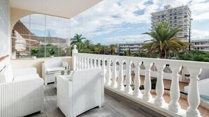 Un balcon doté de balustrades et de mobilier blancs, offrant une vue sur un paysage urbain avec des immeubles et des palmiers.