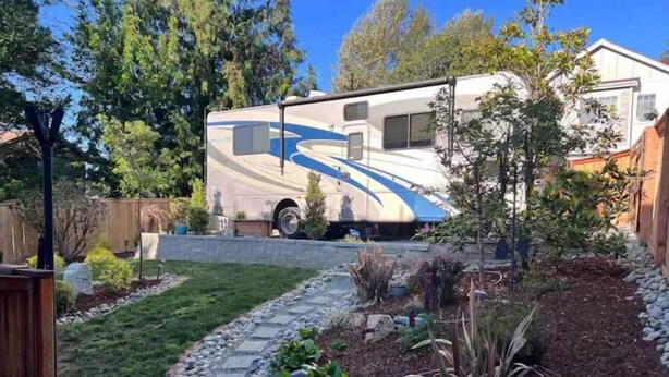 Una caravana estacionada en un patio trasero con un sendero de piedra, varias plantas y una valla de madera.
