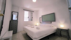 Ein Schlafzimmer mit Bett, Kommode, Schreibtisch, einem Fenster mit Jalousien und einem an der Wand befestigten Fernseher.