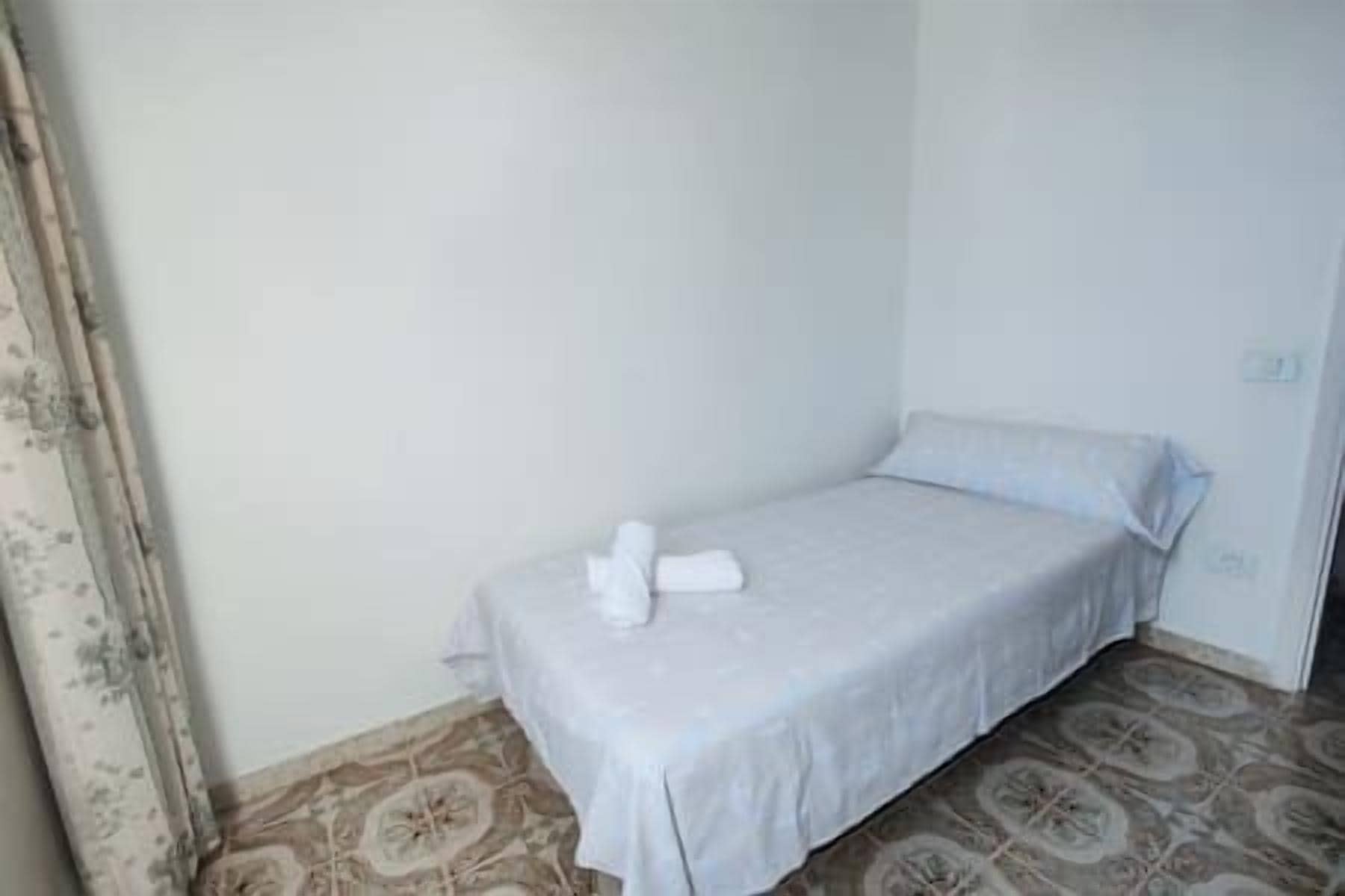 Una habitación pequeña con una cama individual, un edredón blanco sencillo y almohadas, piso estampado y pared lisa.