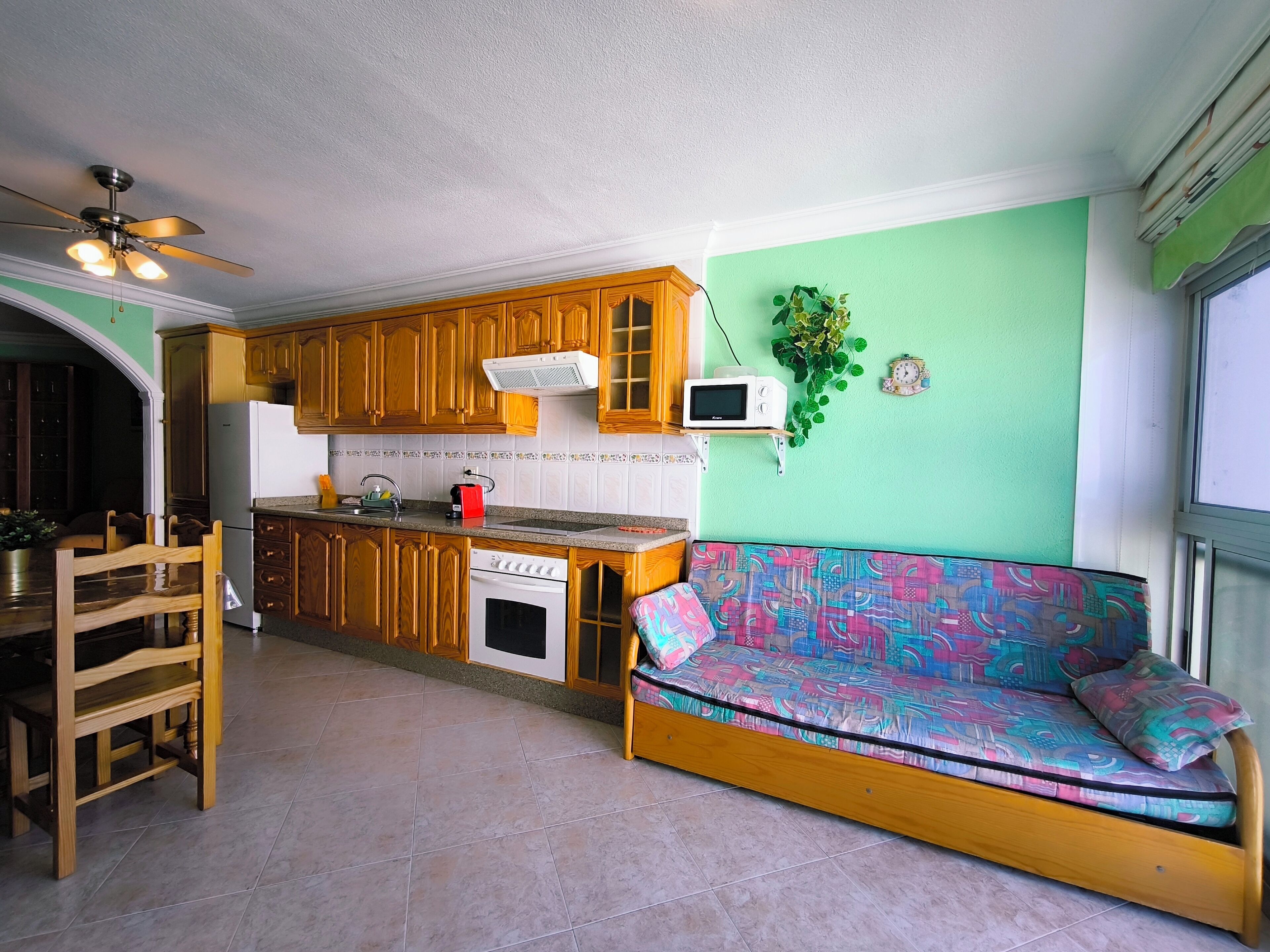 A compact kitchen with wooden cabinets, a microwave, and a built-in oven. A colorful sofa is positioned against the wall, and a ceiling fan is visible.