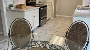 A kitchen with white cabinets, a stove, and a dining area with wicker chairs and a glass table. - Warm welcoming stay (Mandeville)