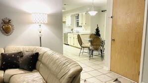 A living room with a beige sofa, a lamp, and a decorative item on the wall. There is a dining area with a table and chairs, and a kitchen in the background. - Warm welcoming stay (Mandeville)