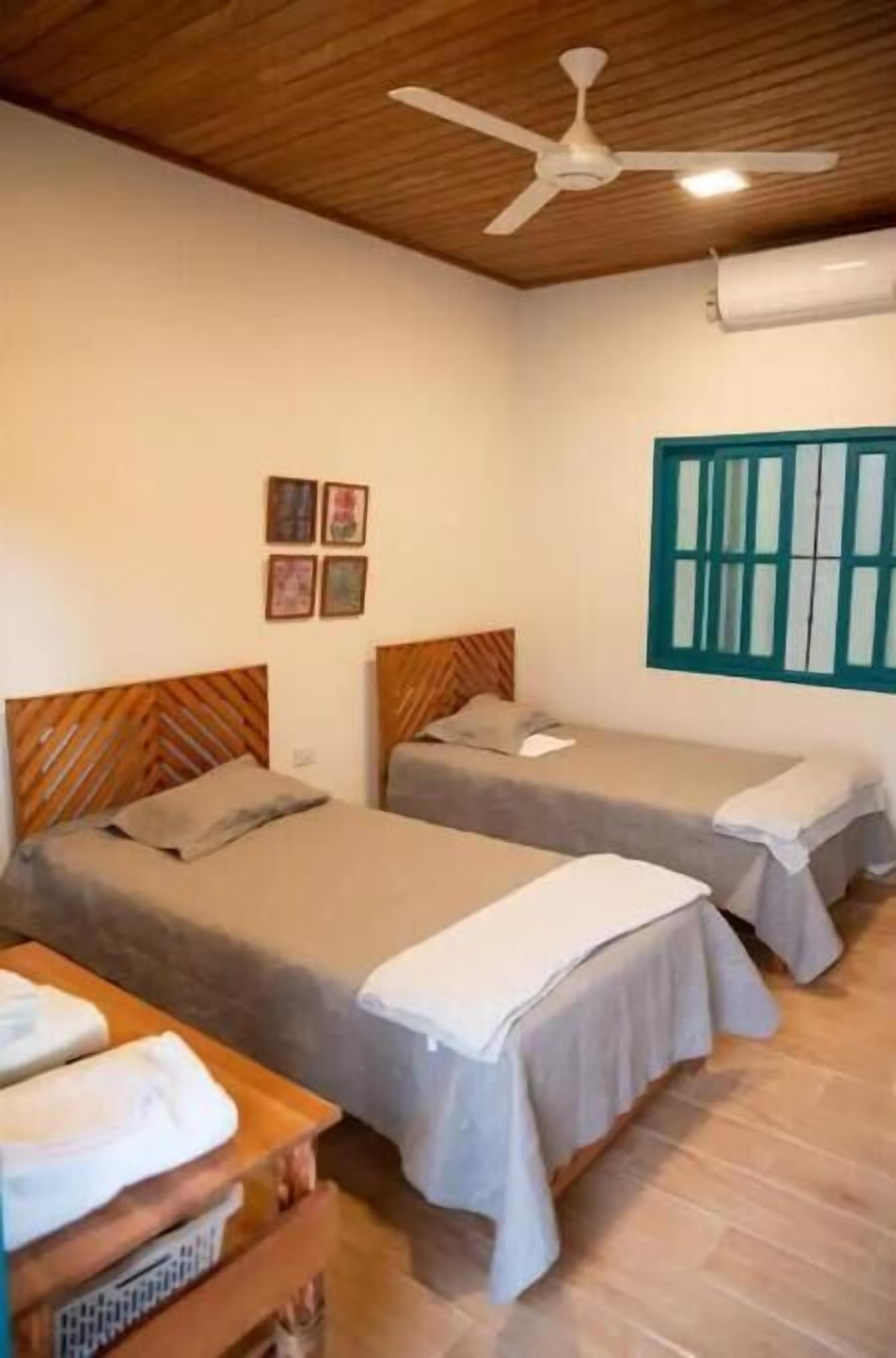 A room with two beds, a ceiling fan, a window with blue shutters, and a wall-mounted air conditioner.