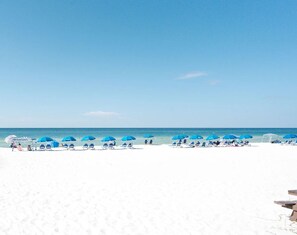 Une plage de sable blanc, sous un ciel d’un bleu limpide. On y voit des rangées de parasols bleus et de chaises longues blanches.