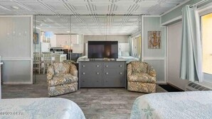 Hotelkamer met een grote televisie, twee gestoffeerde fauteuils, een centraal meubelstuk en uitzicht op de keuken en eetruimte.