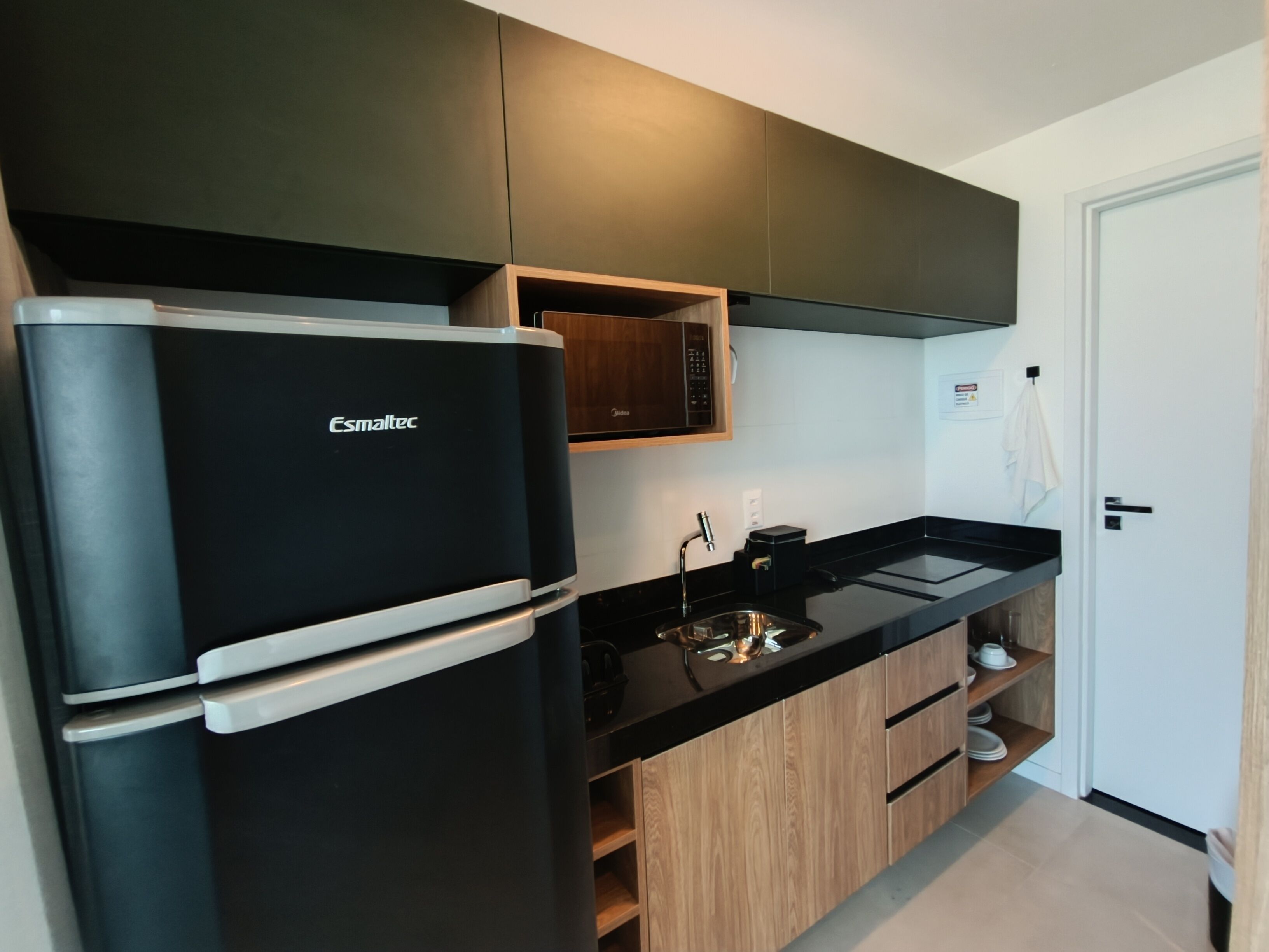 A modern kitchen with a black Esmaltec refrigerator, wooden cabinets, and a black countertop.