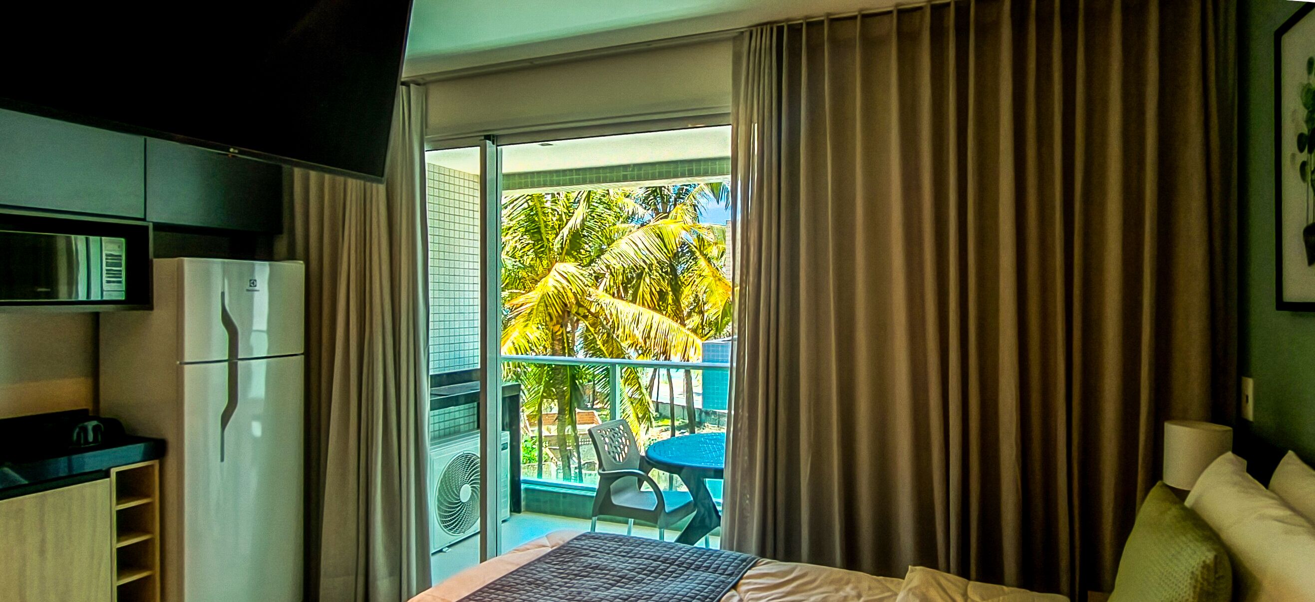 A hotel room with a bed, a small table, a chair, a refrigerator, and a sliding glass door leading to a balcony with a view of palm trees.