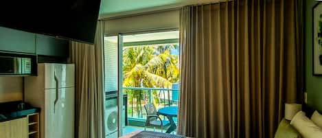 A hotel room with a bed, a small table, a chair, a refrigerator, and a sliding glass door leading to a balcony with a view of palm trees.