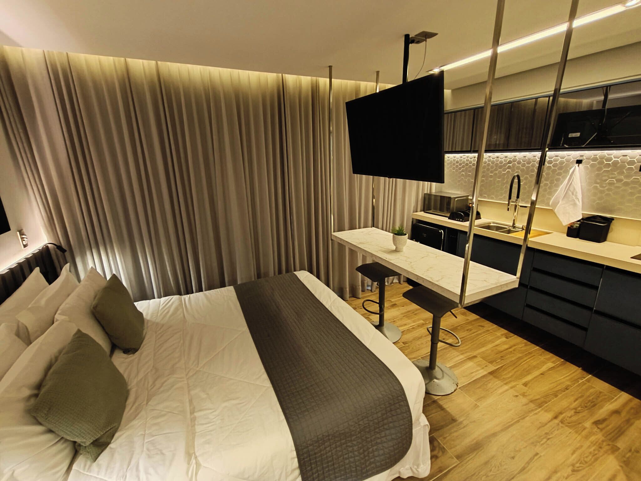 A modern hotel room with a large bed, a desk, and a kitchen area.