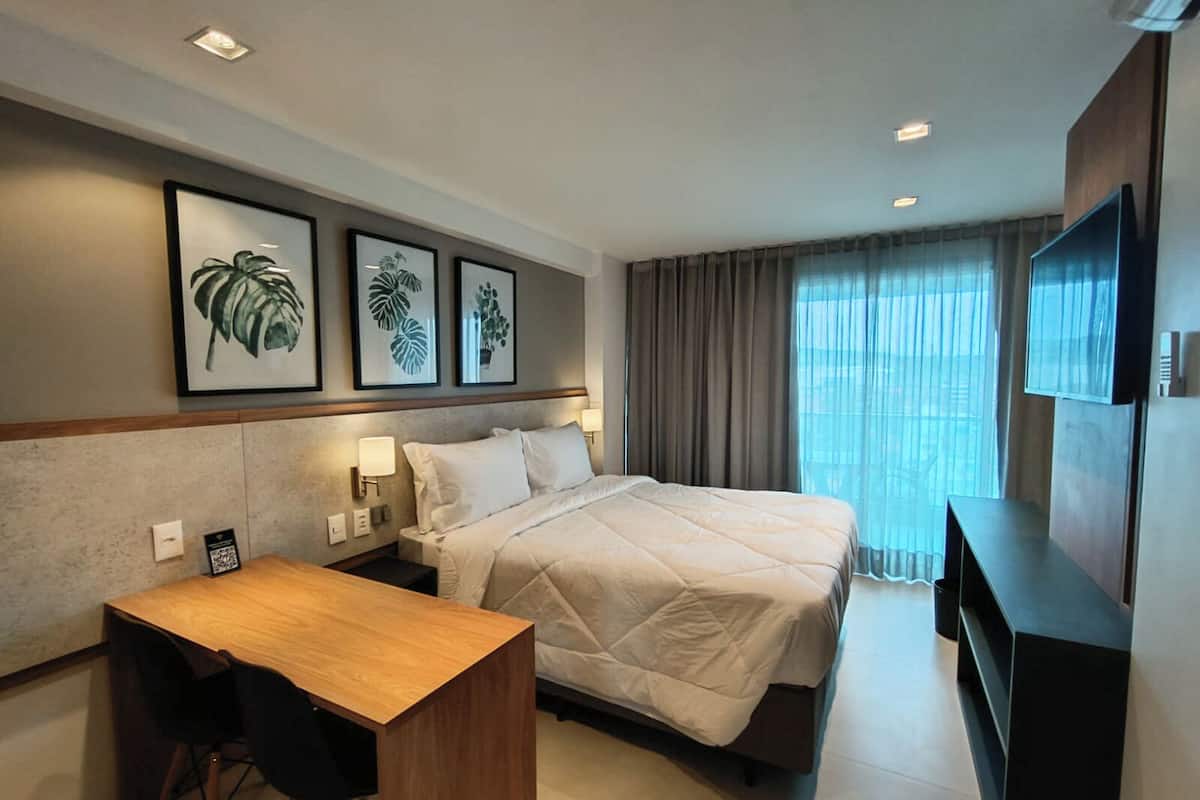 A hotel room with a bed, a desk, a chair, a TV, and framed plant art on the wall.