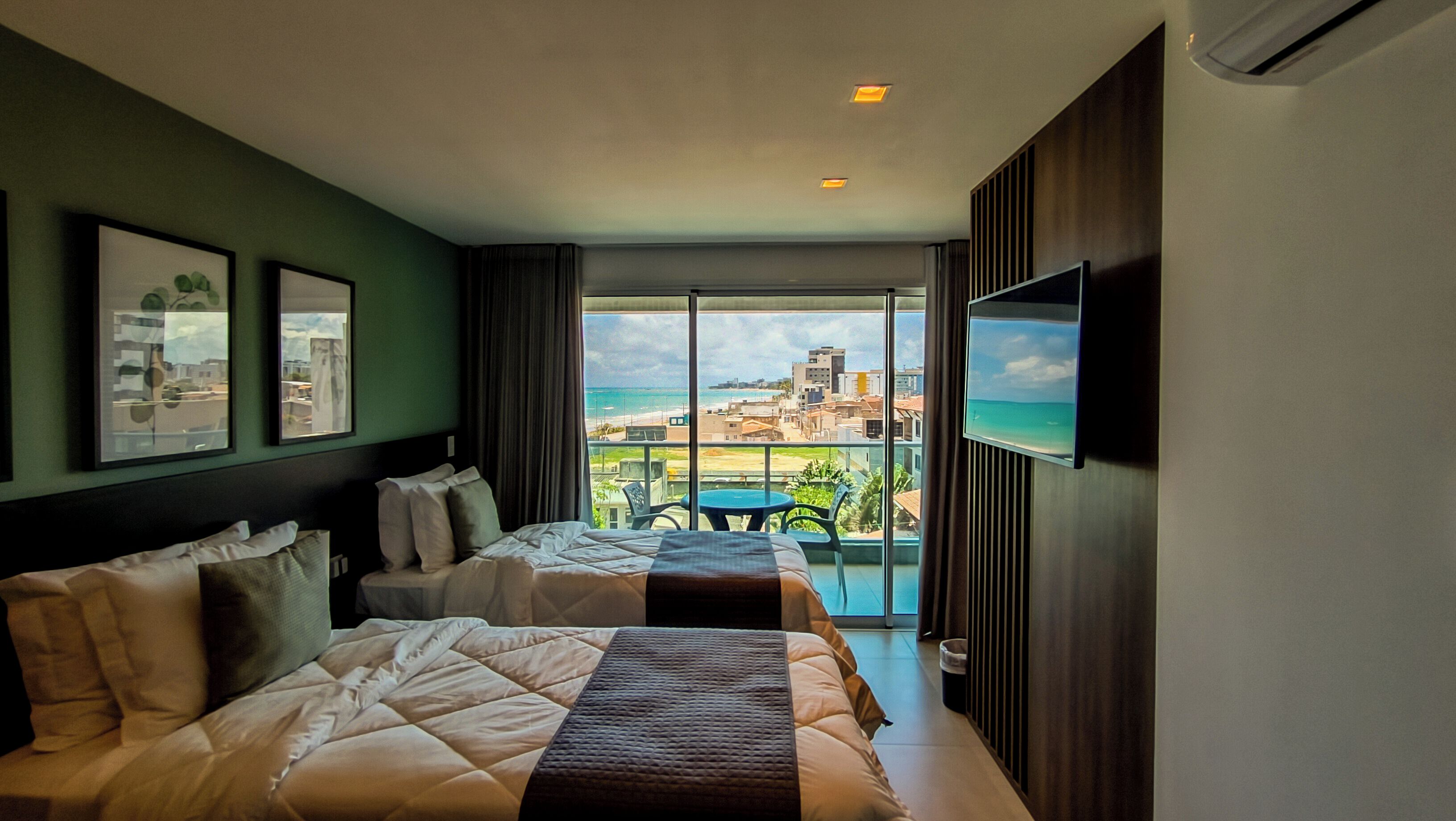 A hotel room with two beds, a TV, and a balcony with a view of the beach and cityscape.