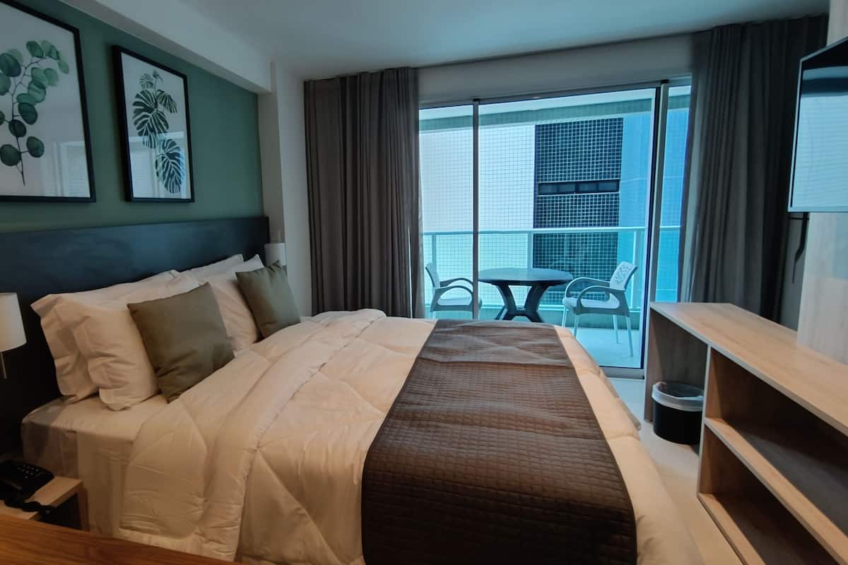 A hotel room with a bed, a desk, a chair, a table, and a balcony with a view of a building.