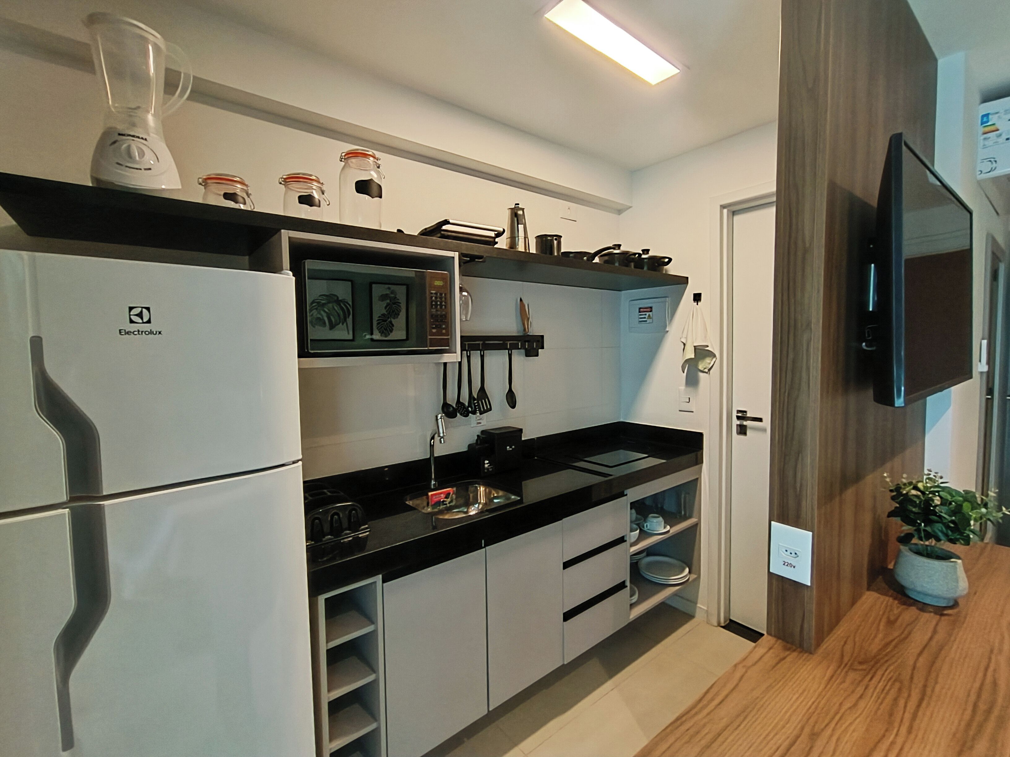 Een moderne keuken met een koelkast, magnetron en fornuis. Er is een plank met potten en een plant.