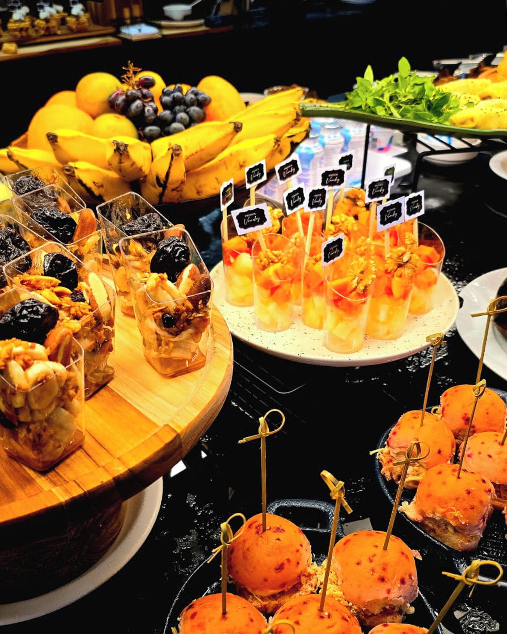 Buffet com diversos alimentos, incluindo frutas, sobremesas e petiscos.