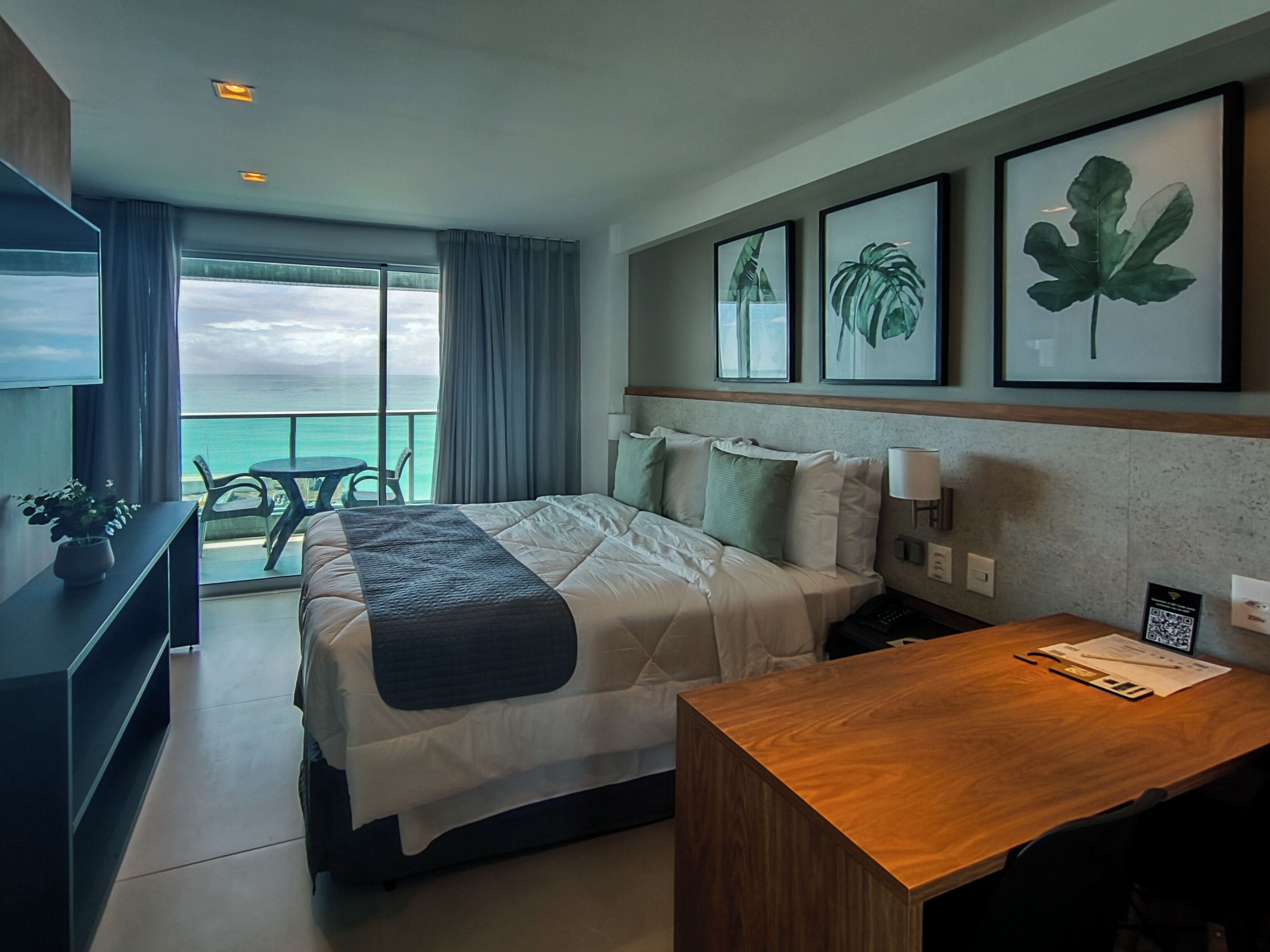 A hotel room with a bed, a desk, a chair, a TV, and a balcony with a view of the ocean.