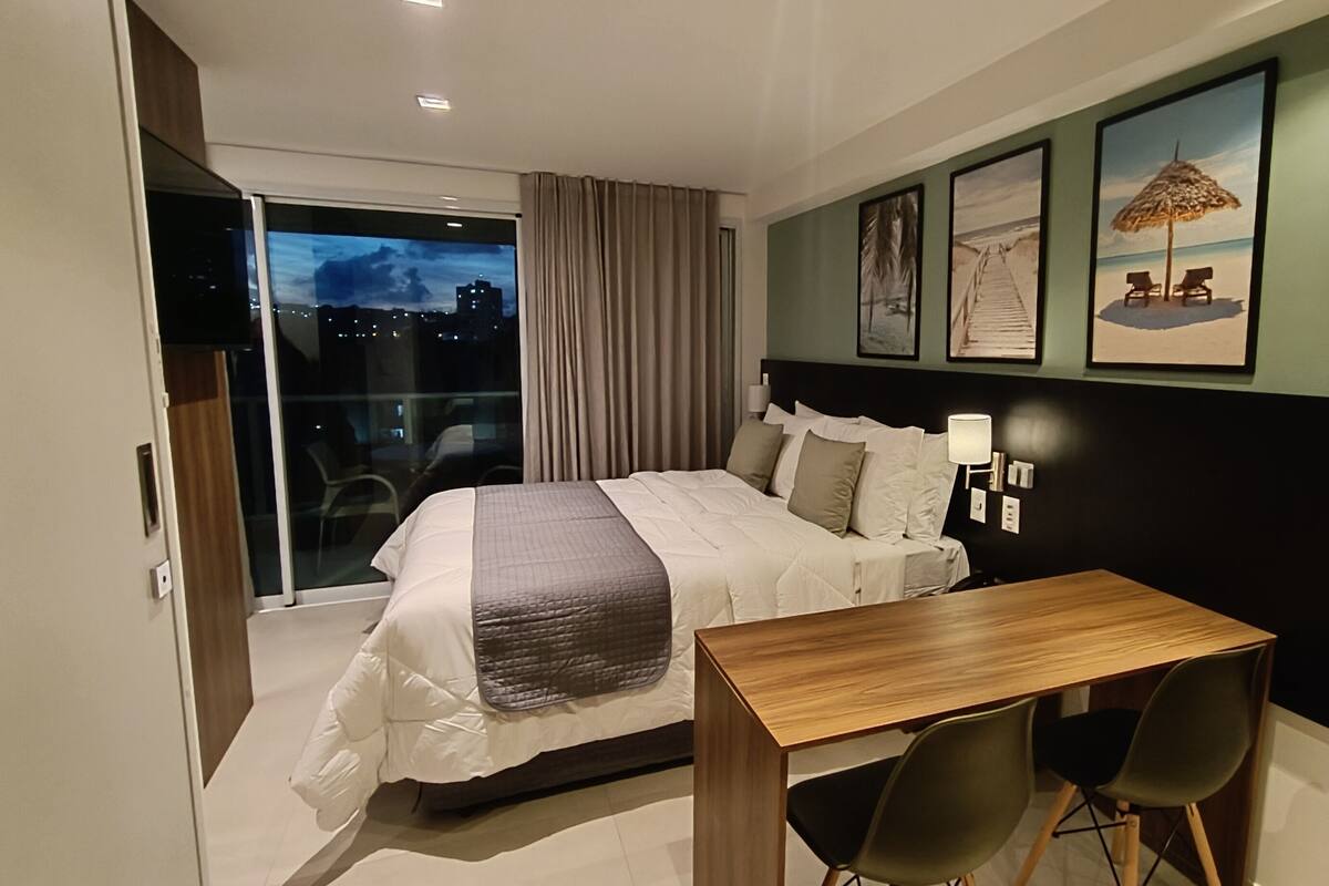 A hotel room with a bed, a desk, and chairs. There is a window with curtains, a wall with framed pictures, and a night view outside.