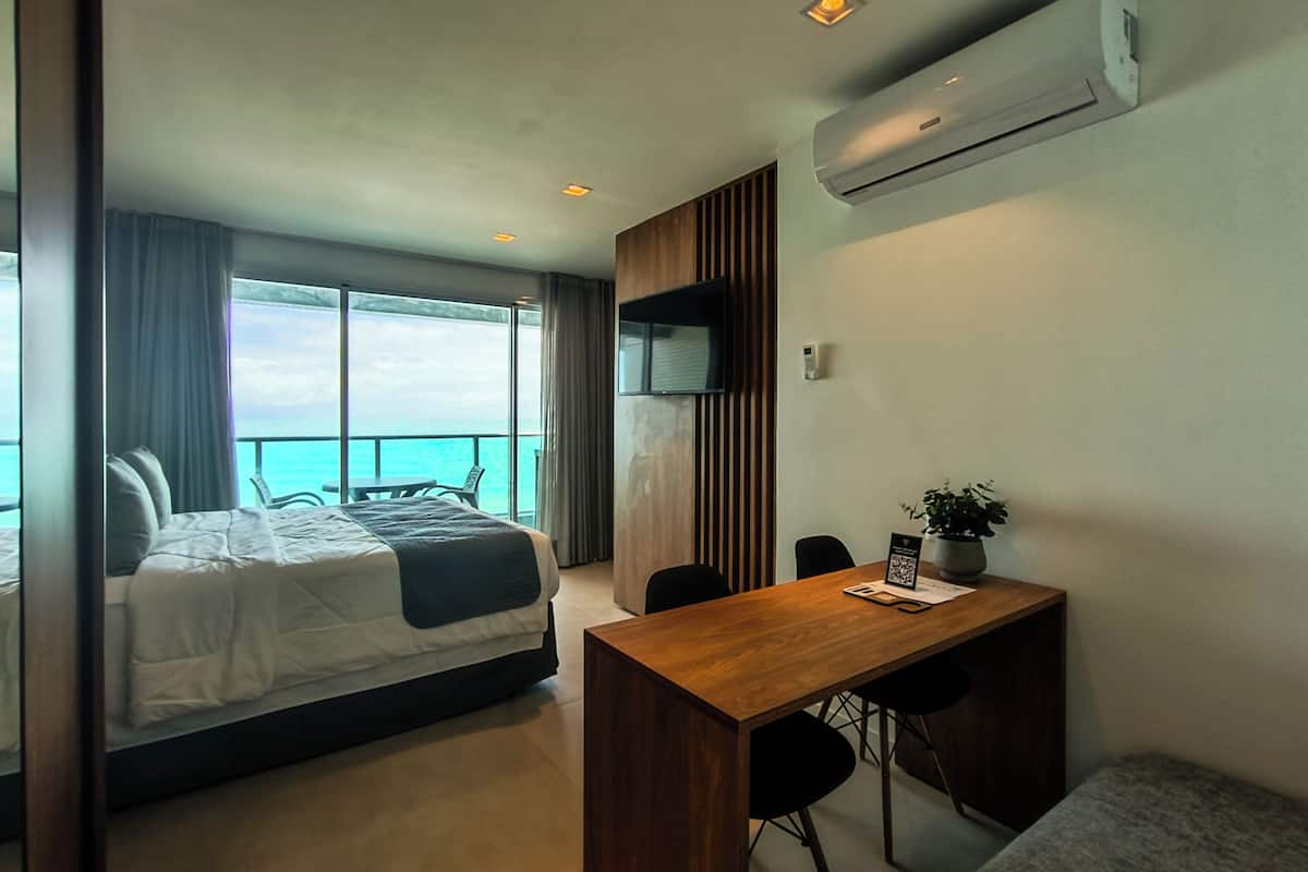 A modern hotel room with a bed, a desk, and a view of the sea.