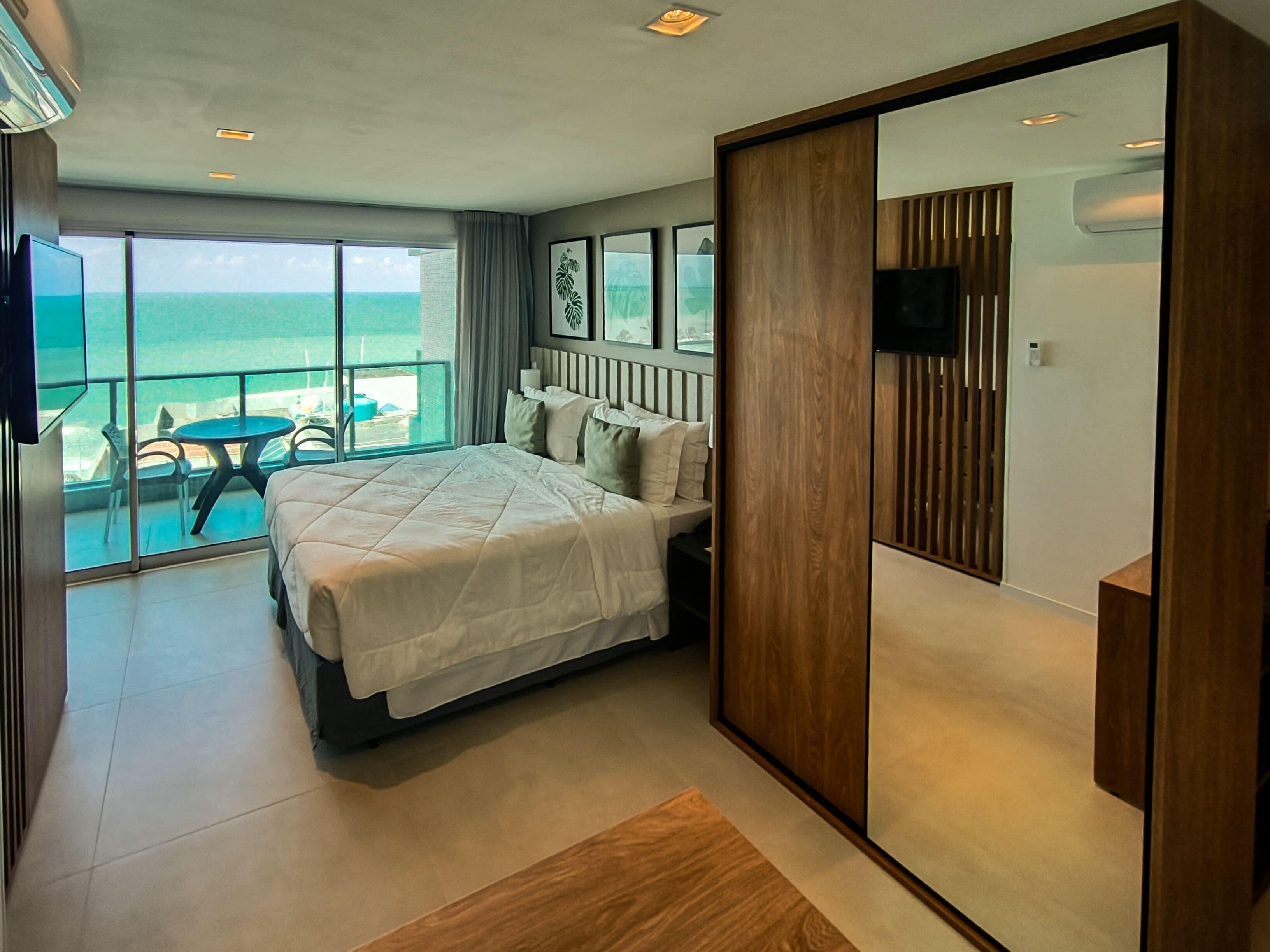 A modern hotel room with a large bed, a balcony with a view of the ocean, and a TV mounted on the wall.
