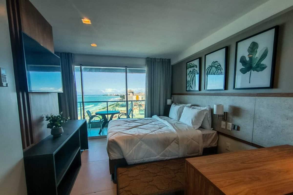 A hotel room with a bed, a desk, a TV, and a balcony with a view of the beach.