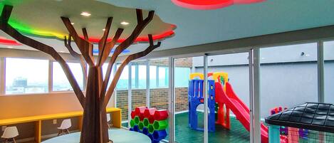 Un espace de jeux intérieur moderne, doté d’une structure colorée, d’une piscine à balles et d’une grande sculpture en forme d’arbre.