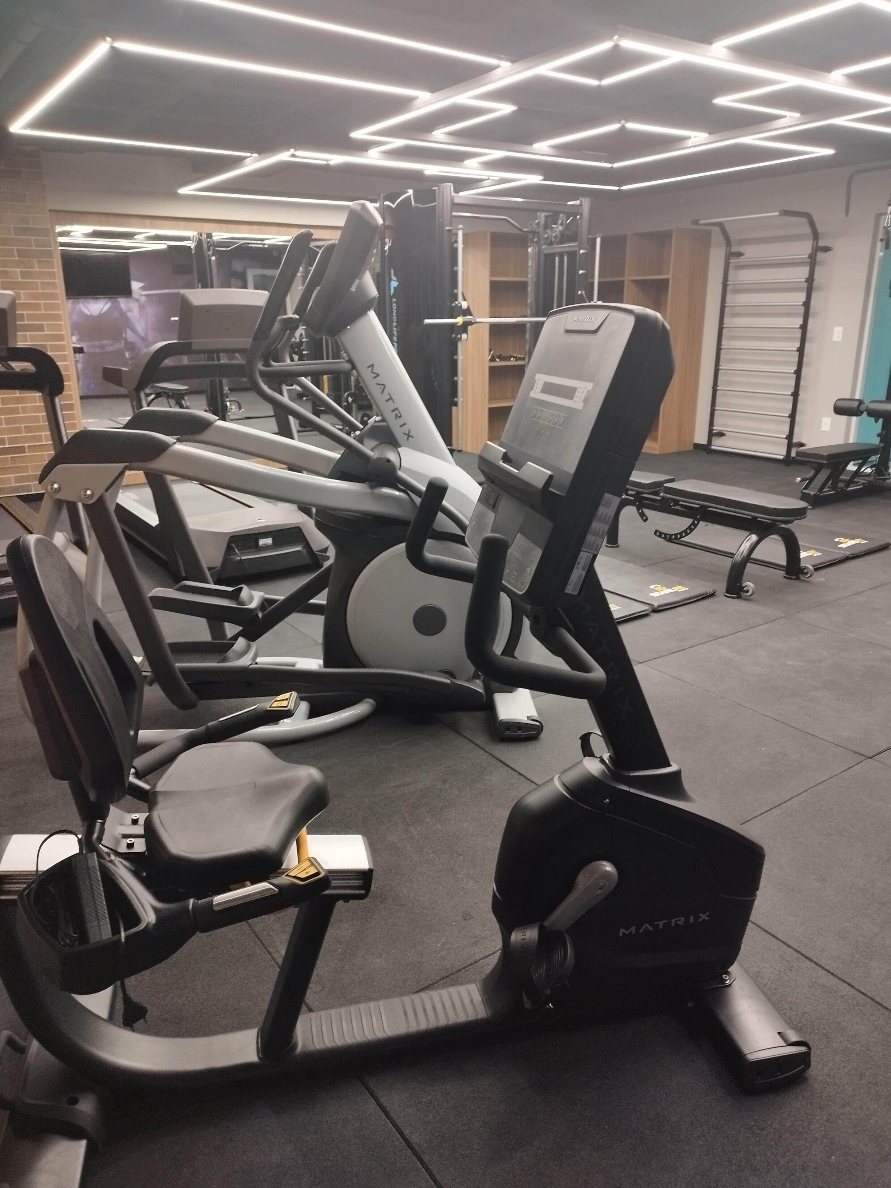 Un gimnasio con diverso equipo de ejercicio, incluyendo cintas de correr y máquinas de pesas, bajo una iluminación moderna.