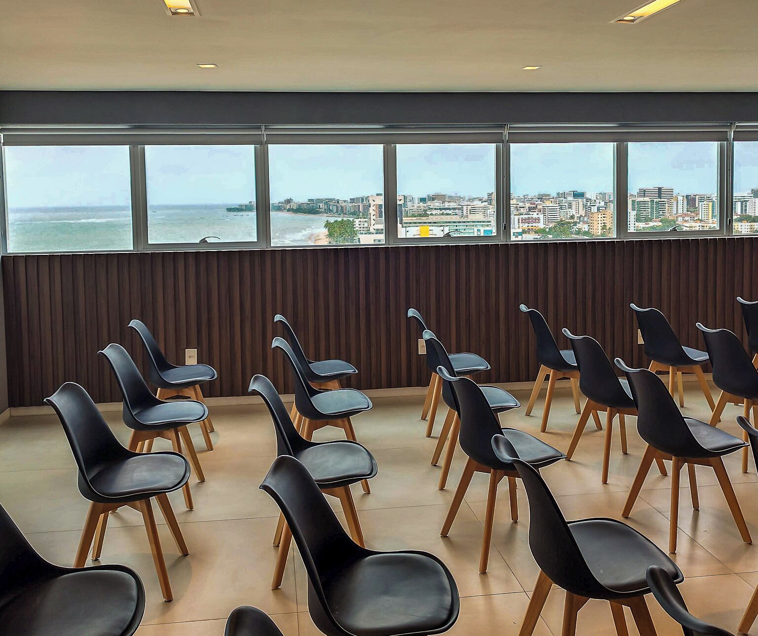 Un salón de conferencias con filas de sillas negras de patas de madera, ventanas amplias con vista a la ciudad y un panel de pared de madera.