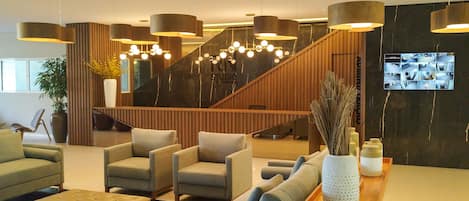 A modern lobby with sofas, armchairs, a wooden staircase, and a reception desk.