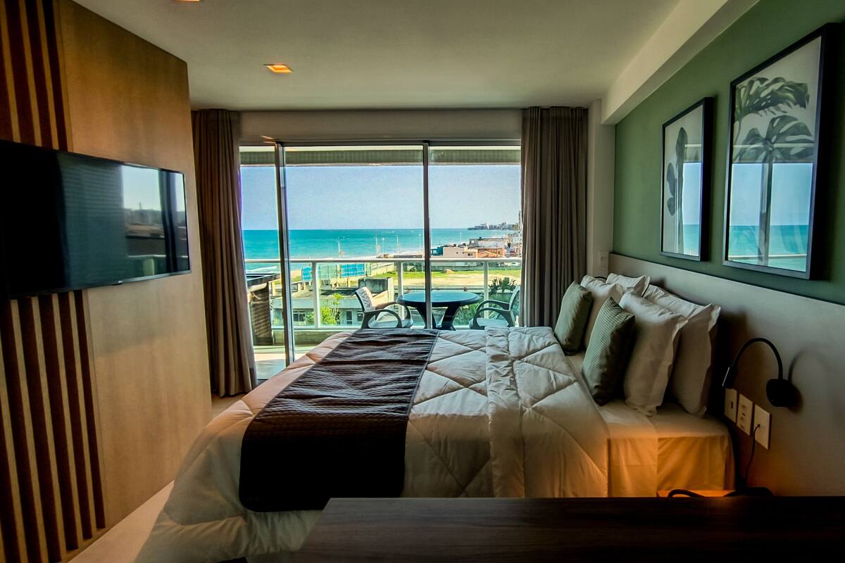 A hotel room with a bed, a TV, a large window with a view of the beach and cityscape, and a table with chairs.