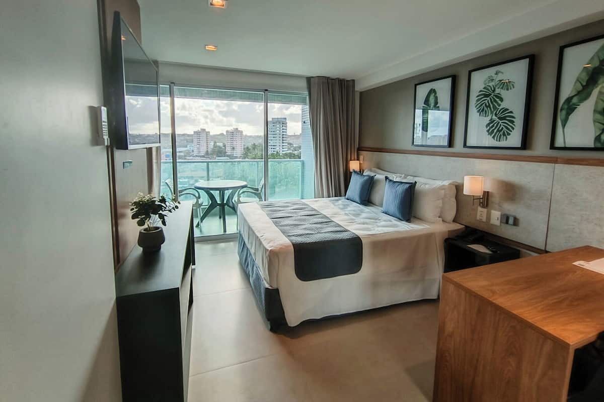 A modern hotel room with a large bed, bedside tables, a small desk, and a balcony with a view of the city.