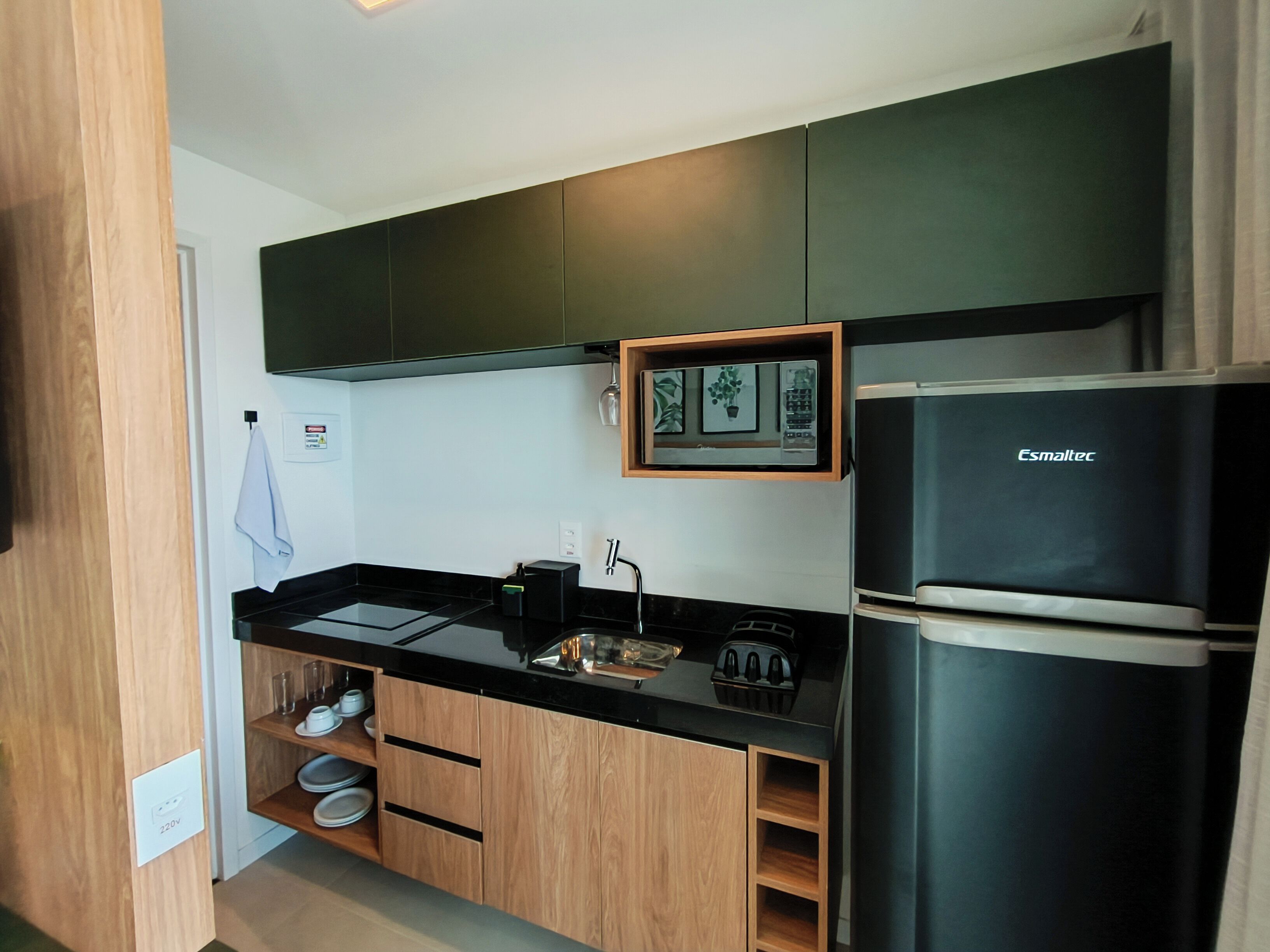 A modern kitchen with black countertops, wooden cabinets, and a black Esmaltec refrigerator.