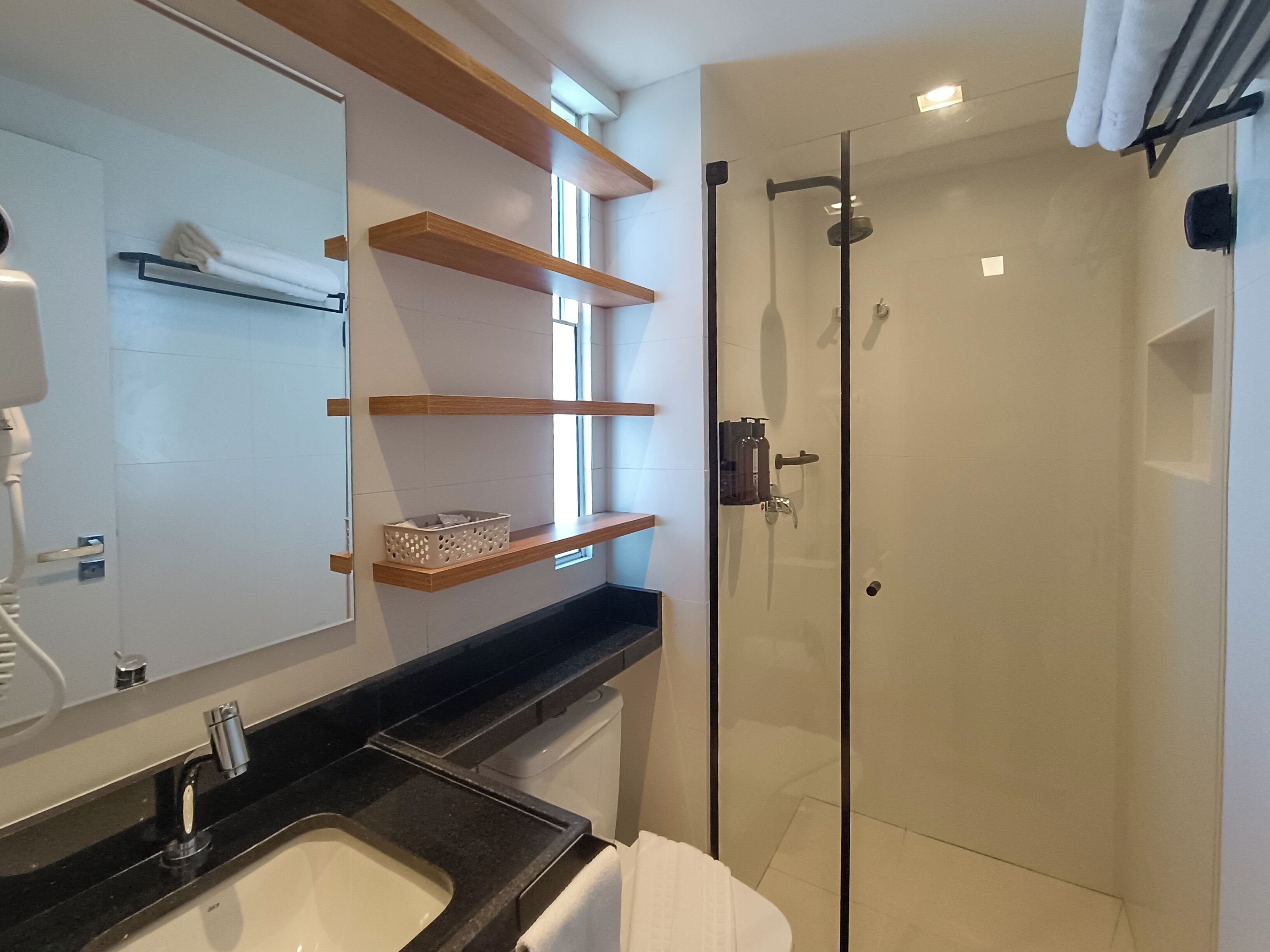 A modern bathroom with a glass shower enclosure, a sink with a black countertop, and wooden shelves above the mirror.
