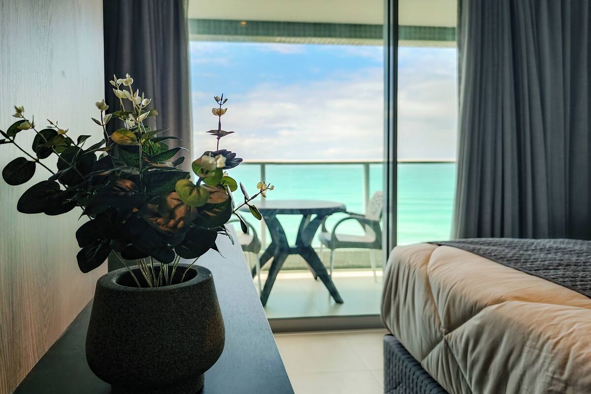 A room with a bed, a potted plant, a table, and a view of the ocean.