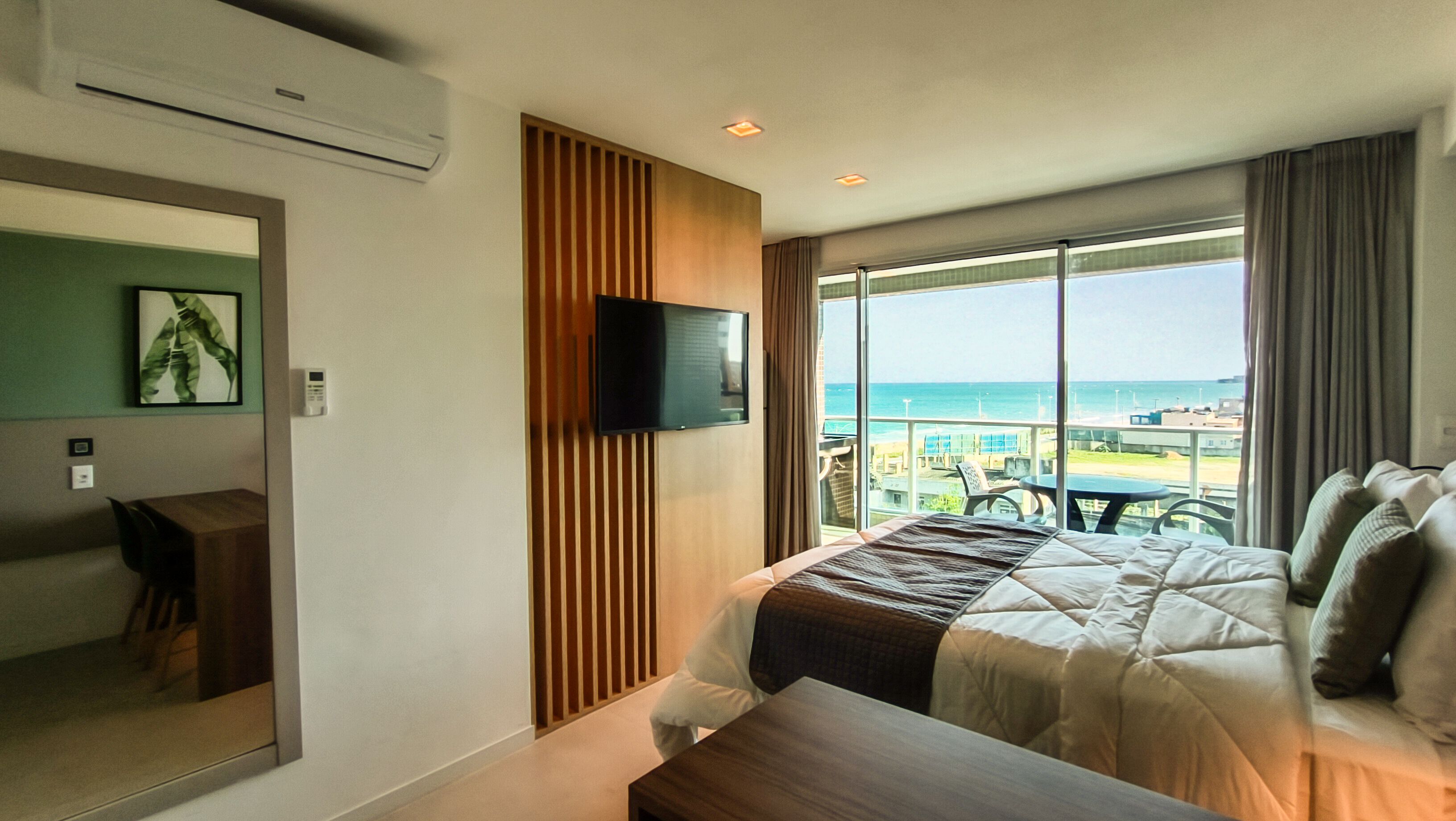 A hotel room with a bed, a TV, a desk, and a view of the beach.