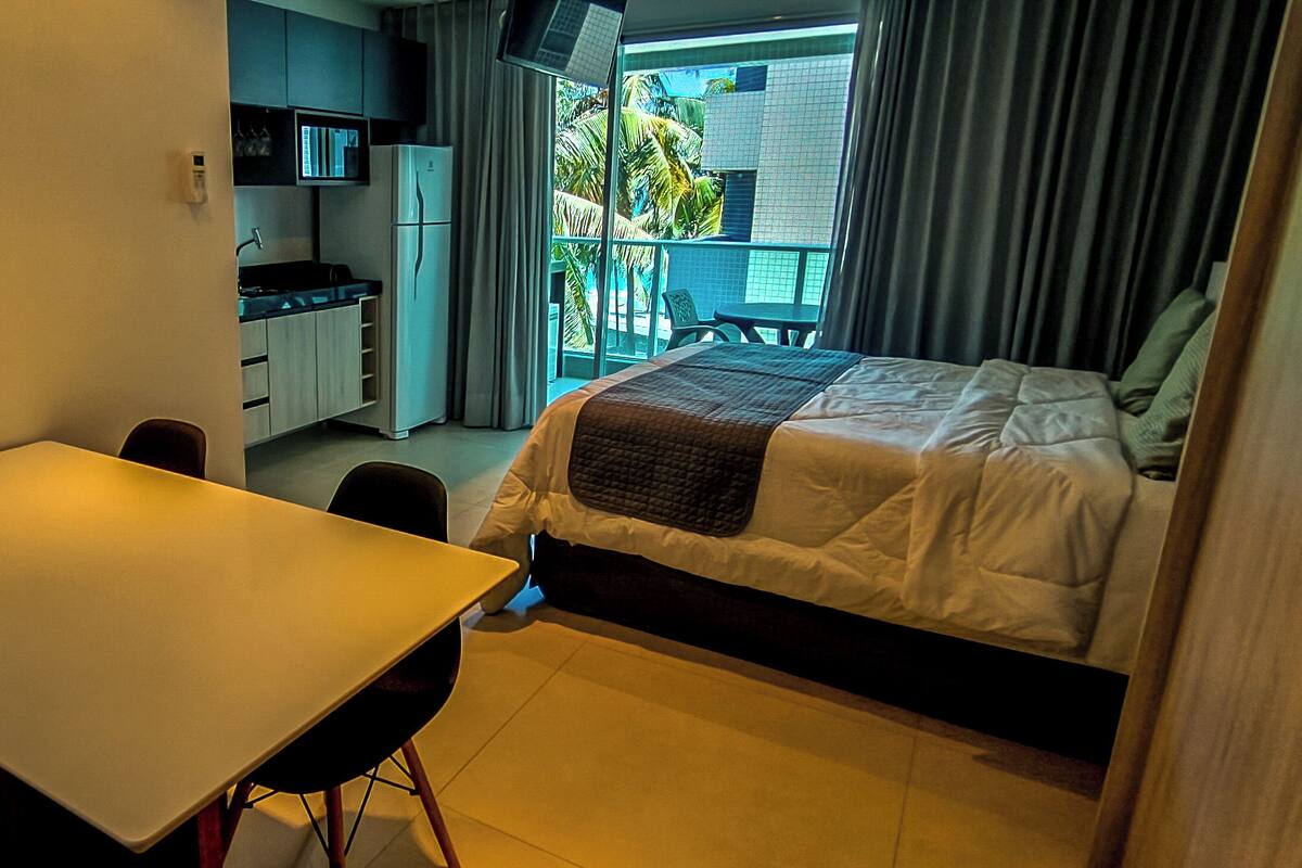 A hotel room with a bed, a desk, a chair, a small kitchenette, and a balcony with a view of palm trees.