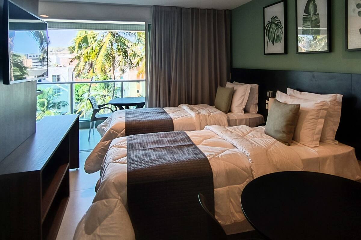 A hotel room with two beds, a desk, and a chair. There are framed pictures on the wall and a view of palm trees outside.