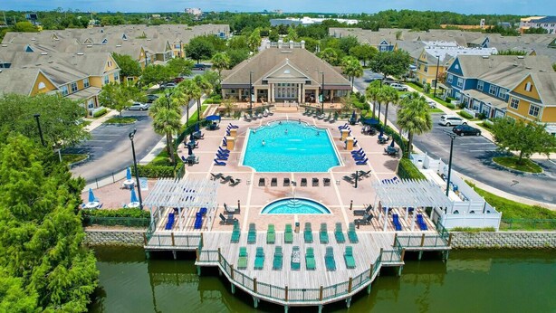 A large swimming pool surrounded by lounge chairs and a dock extending into a body of water, with residential buildings in the background. - Vilas em Seven Dwarfs - Orla1 (Kissimmee)