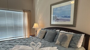 A neatly made bed with a grey quilt, a pair of white towels arranged in a bow, a lamp on a bedside table, and a framed picture of a beach scene on the wall. - Vilas em Seven Dwarfs - Orla1 (Kissimmee)