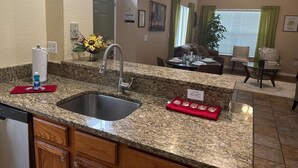 A kitchen with granite countertops, a stainless steel sink, wooden cabinets, and a dining area with a table set for four. - Vilas em Seven Dwarfs - Orla1 (Kissimmee)