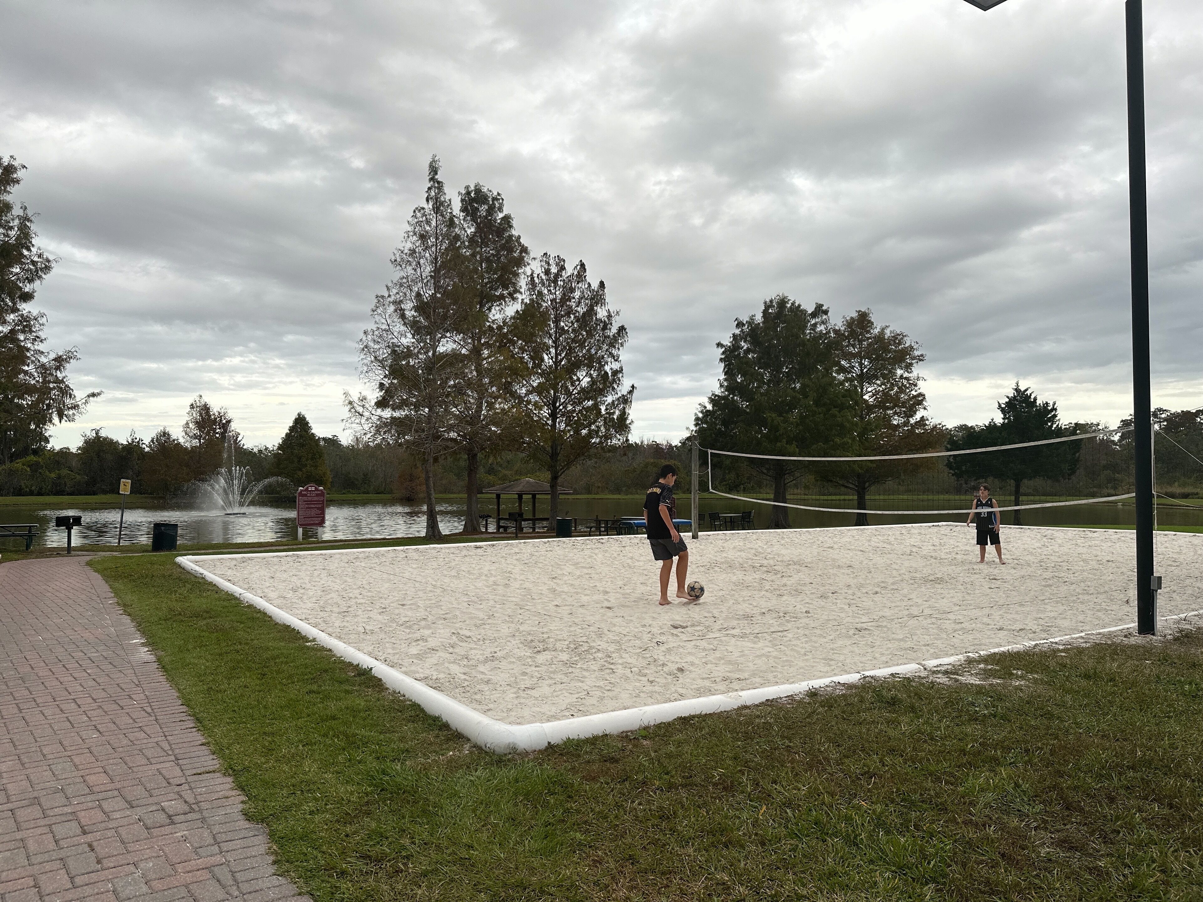 A volleyball court with a sandy surface, surrounded by a grassy area and trees.