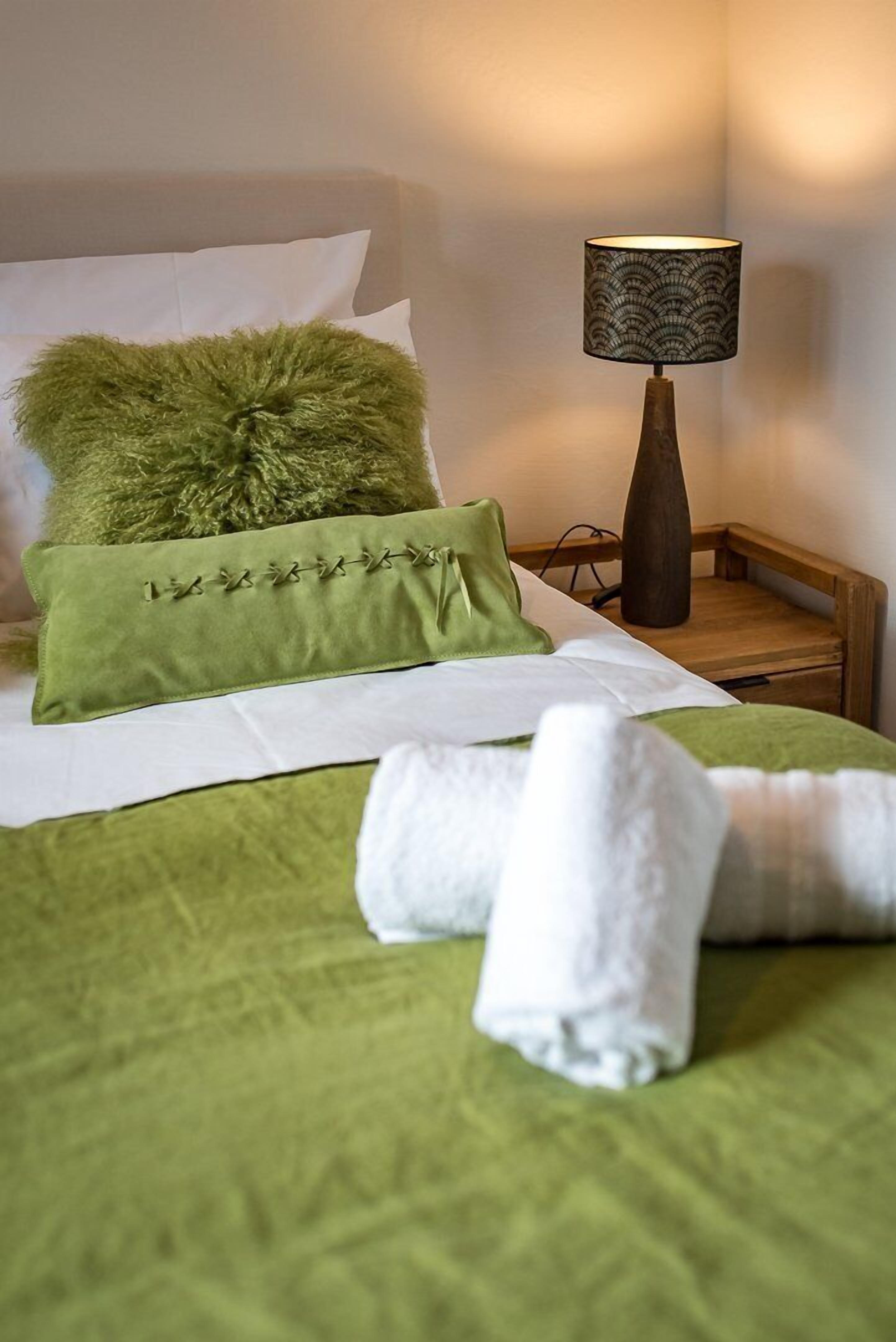 A neatly made bed with green bedding, a fluffy green pillow, and a rolled white towel on the bed.