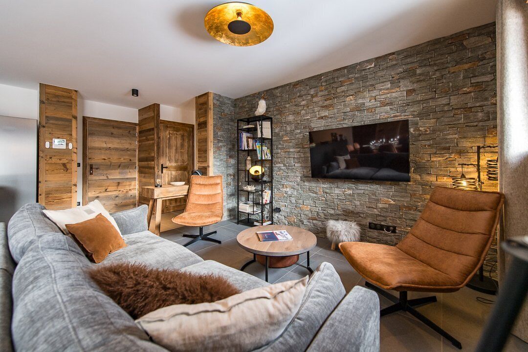 Een moderne woonkamer met een stenen muur, een flatscreen televisie, een grijze bank en een bruine leren fauteuil.