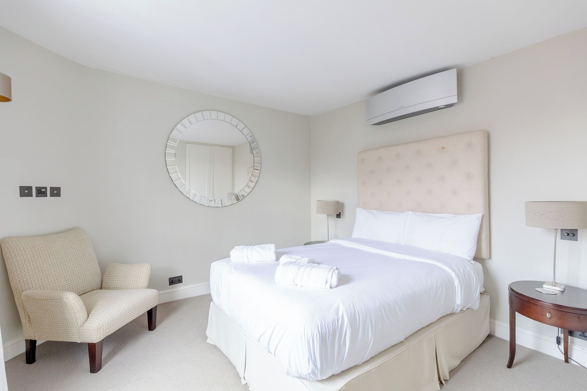 A hotel room with a large bed, a round mirror, a headboard, a chair, a bedside table, and an air conditioning unit.